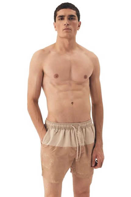 Homme portant un short de bain beige avec des motifs tropicaux subtils, inspiré du style décontracté et festif des plages brésiliennes, idéal pour une promenade en bord de mer ou un moment de détente.