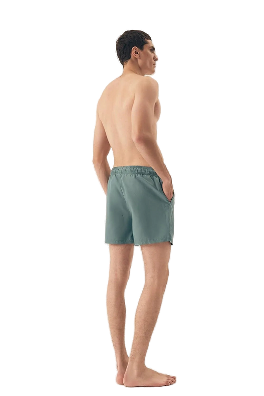 Homme portant un maillot de bain vert au design minimaliste, inspiré de l'esprit détendu et vibrant du Brésil, photographié de dos dans un décor épuré. Idéal pour les journées ensoleillées à la plage ou les activités sportives, ce short incarne l'élégance décontractée des plages de Rio et Bahia.