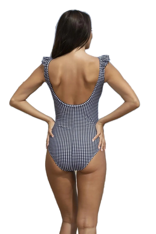 Maillot de bain une-pièce à carreaux noir et blanc avec dos échancré et volants aux épaules, porté sur une plage exotique avec vue sur la mer et des falaises en arrière-plan, incarnant l'élégance brésilienne et l'esprit tropical.